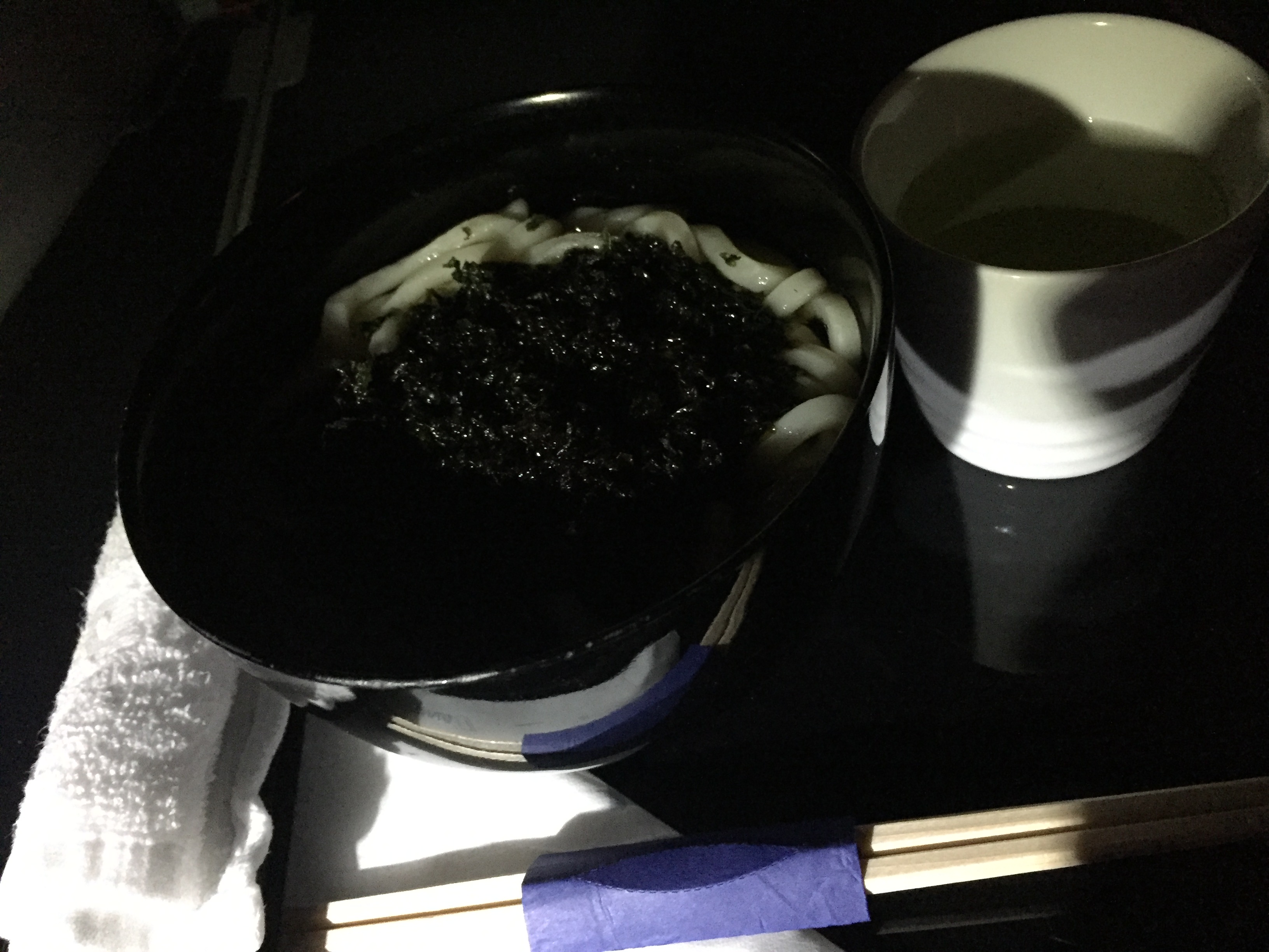A dark bowl of udon noodles topped with a generous mound of nori, served with a small cup of green tea
