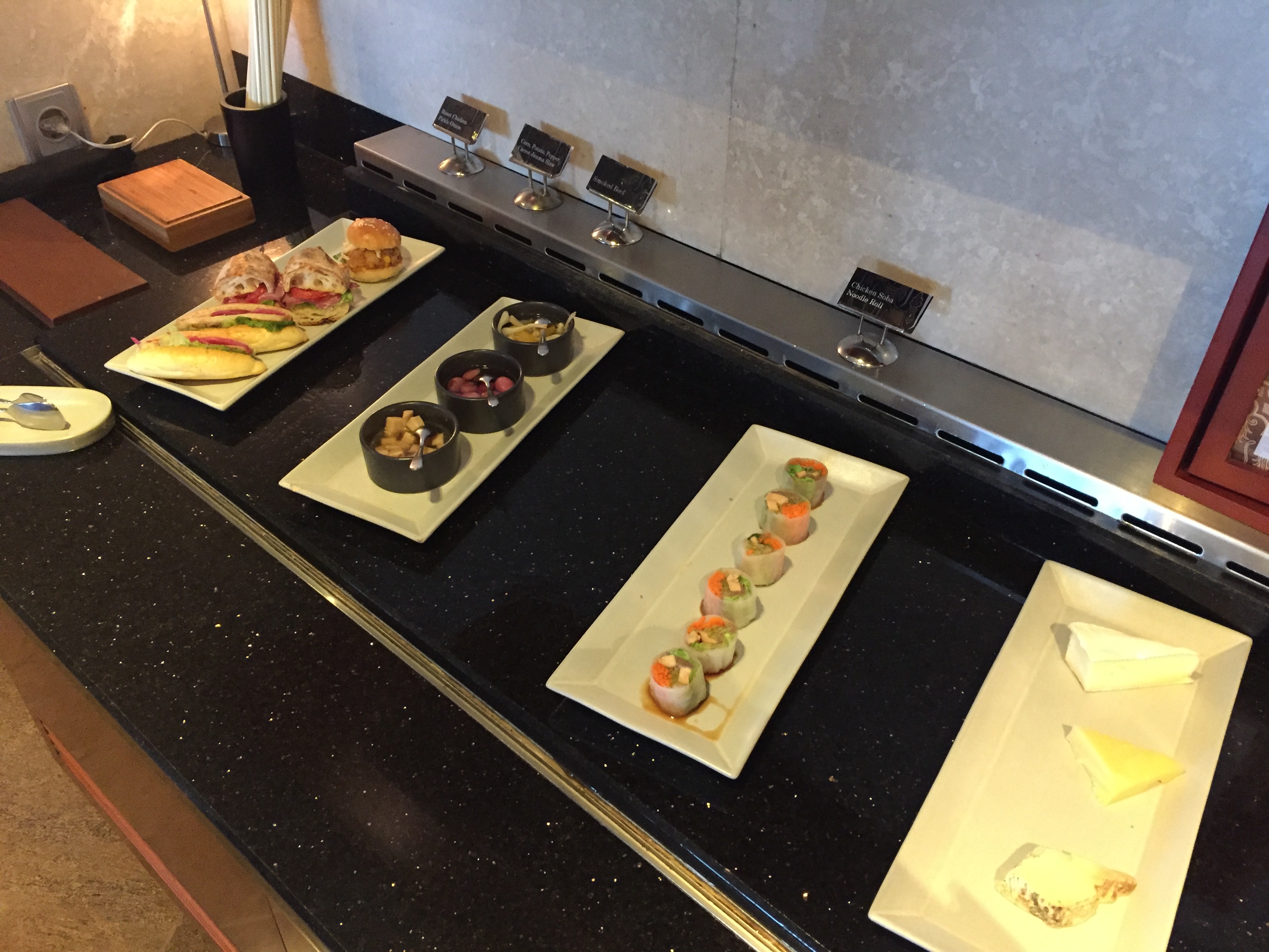 Sandwiches, sushi rolls, salads, and cheese arranged on white rectangular platters along a counter