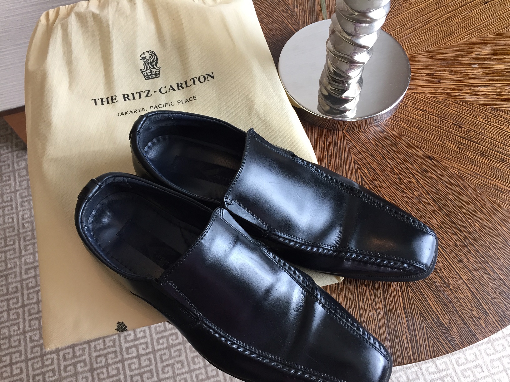 A pair of black leather dress shoes beside a Ritz-Carlton Jakarta, Pacific Place shoe polishing bag on a rattan-and-stone side table