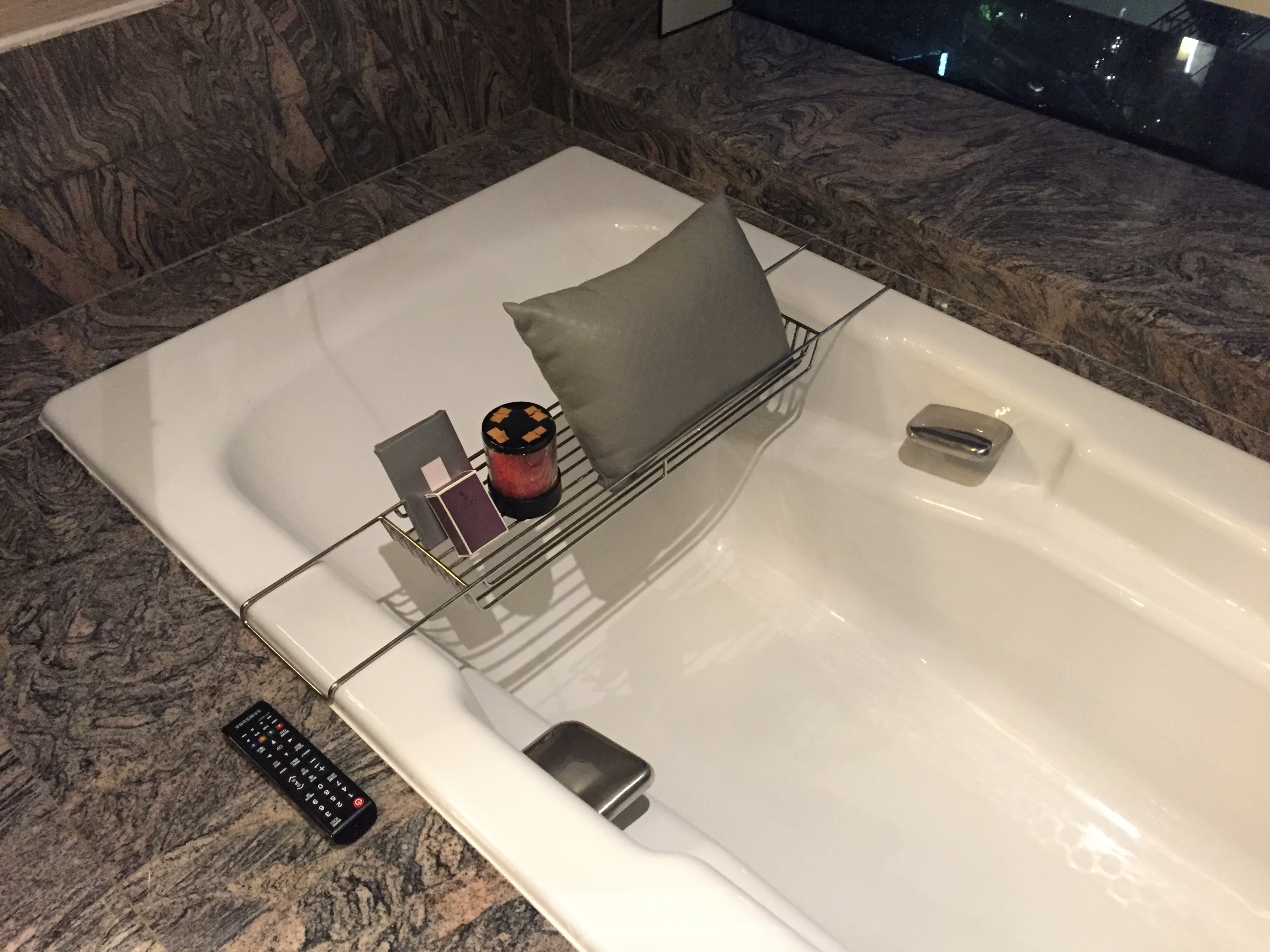 A bath caddy across a deep white tub — grey cushion, candle, small soap or amenity box, and a TV remote on the rim